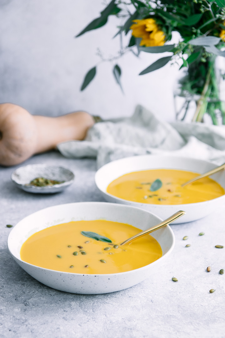 Two white bowls with yellow butternut squash soup and a bouquet of flowers.