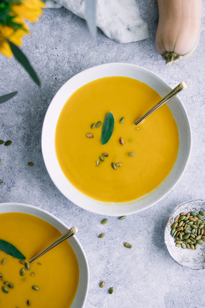 A white bowl with yellow vegan butternut squash soup with a sage leave for garnish.