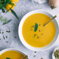 A bowl of yellow butternut squash soup on a blue table with pumpkin seeds.