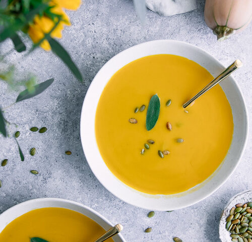 A bowl of yellow butternut squash soup on a blue table with pumpkin seeds.