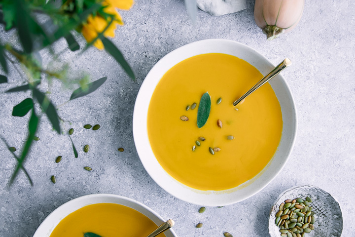 A bowl of yellow butternut squash soup on a blue table with pumpkin seeds.