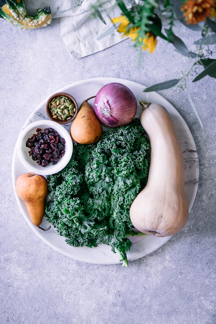 butternut squash, kale, red onion, cranberries, and pumpkin seeds on white cutting board