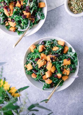 Two white bowls with a fall kale salad on a blue table with orange flowers.
