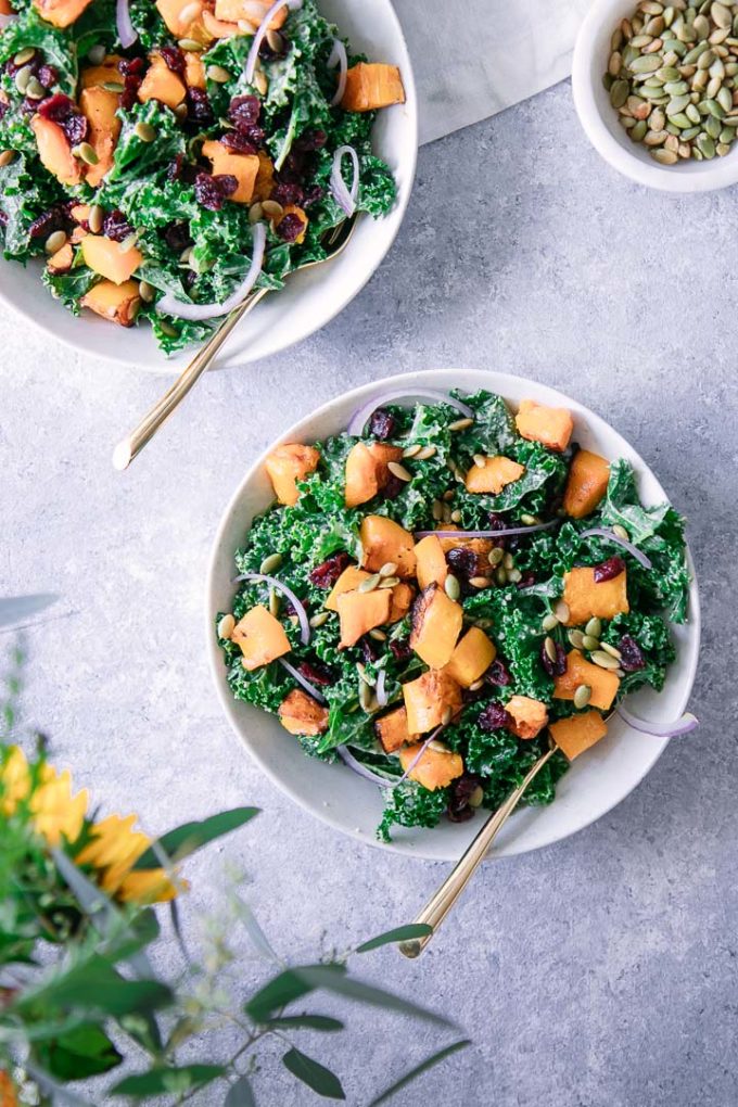 Two white bowls with a fall kale salad on a blue table with orange flowers.