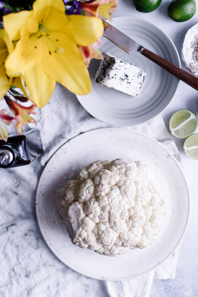 A white plate with a head of cauliflower and a side of blue cheese and cut limes.