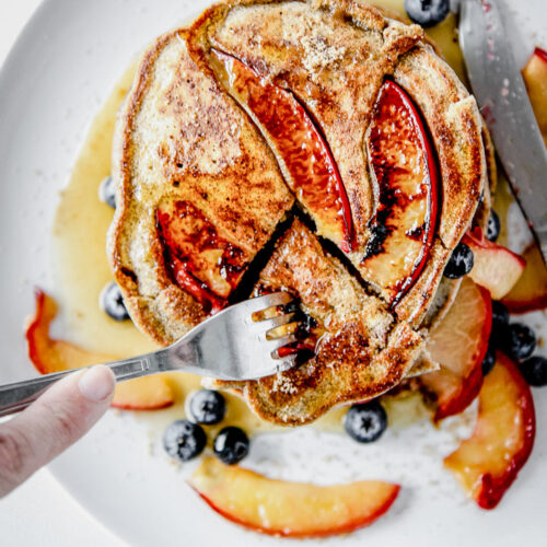 Pluot brown sugar buckwheat pancakes on a white plate being cut with a fork.