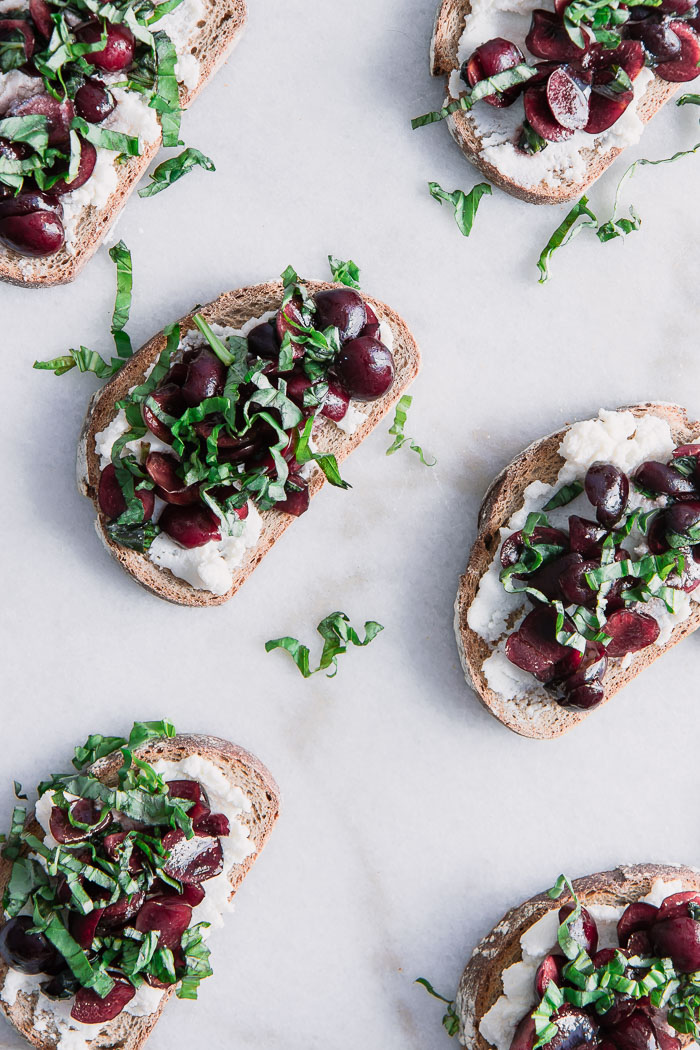 Toast with vanilla ricotta cheese with cherries and basil on top of a white marble table.