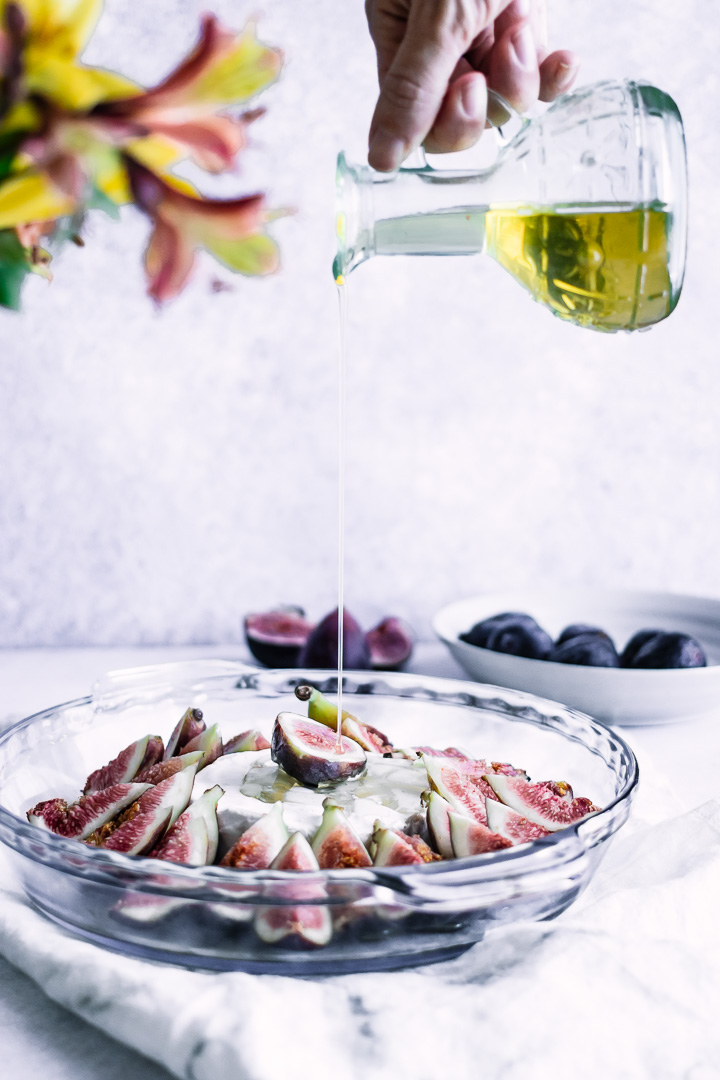 A hand drizzling maple syrup over a glass baking dish with figs and brie.