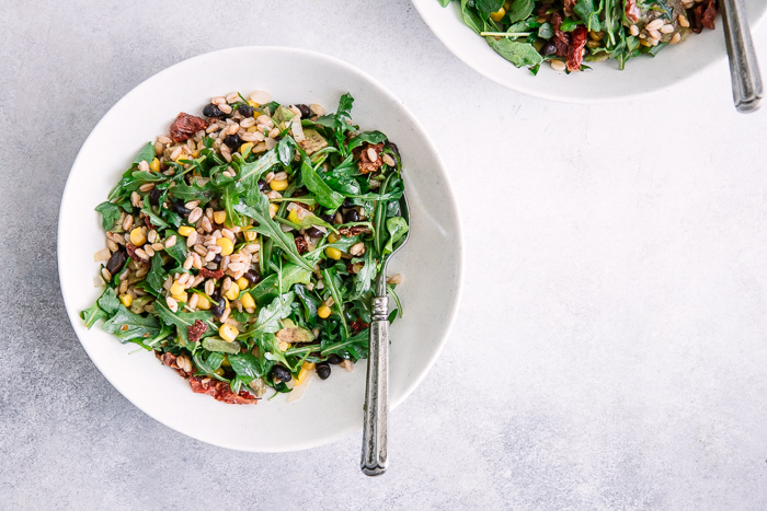 A simple vegan salad with grains, greens, and sun dried tomatoes in two white bowls on a blue table.