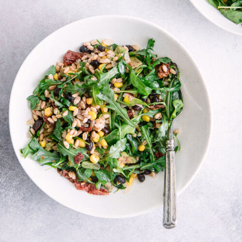 A grain and greens salad in a while bowl on a blue table with a silver fork.