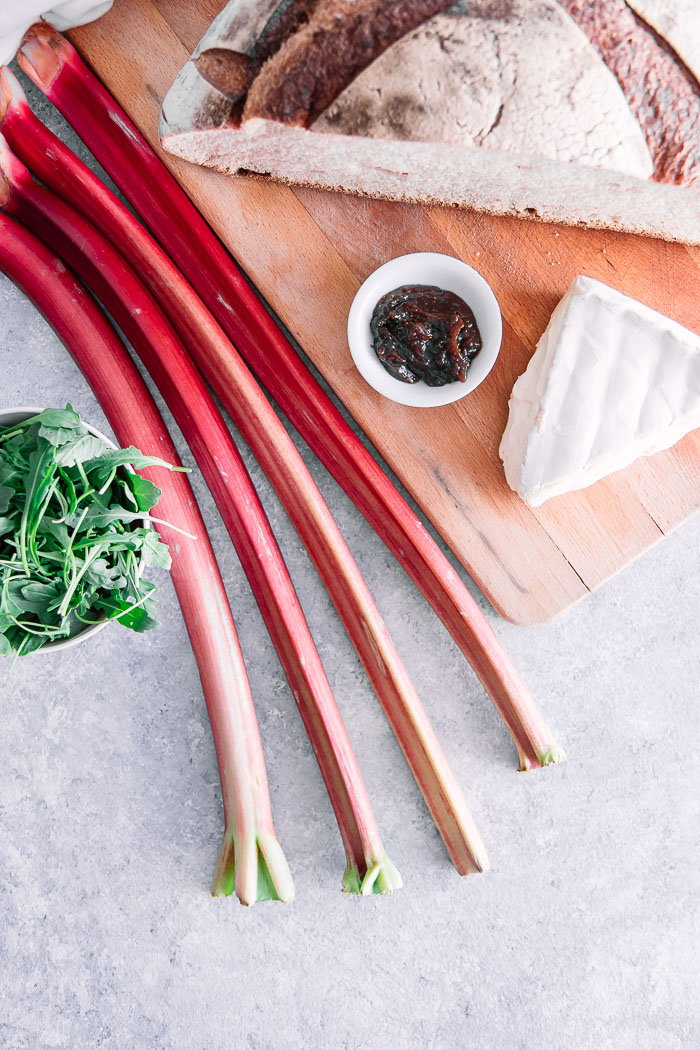 Rhubarb, brie, arugula, jam, and bread on a wooden cutting board.