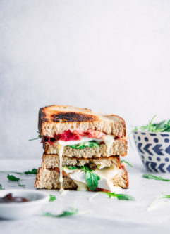 A stacked sandwich with rhubarb, argula, and brie on a white table with jam and a blue bowl.