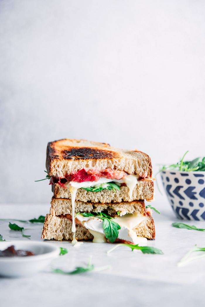 A stacked sandwich with rhubarb, argula, and brie on a white table with jam and a blue bowl.
