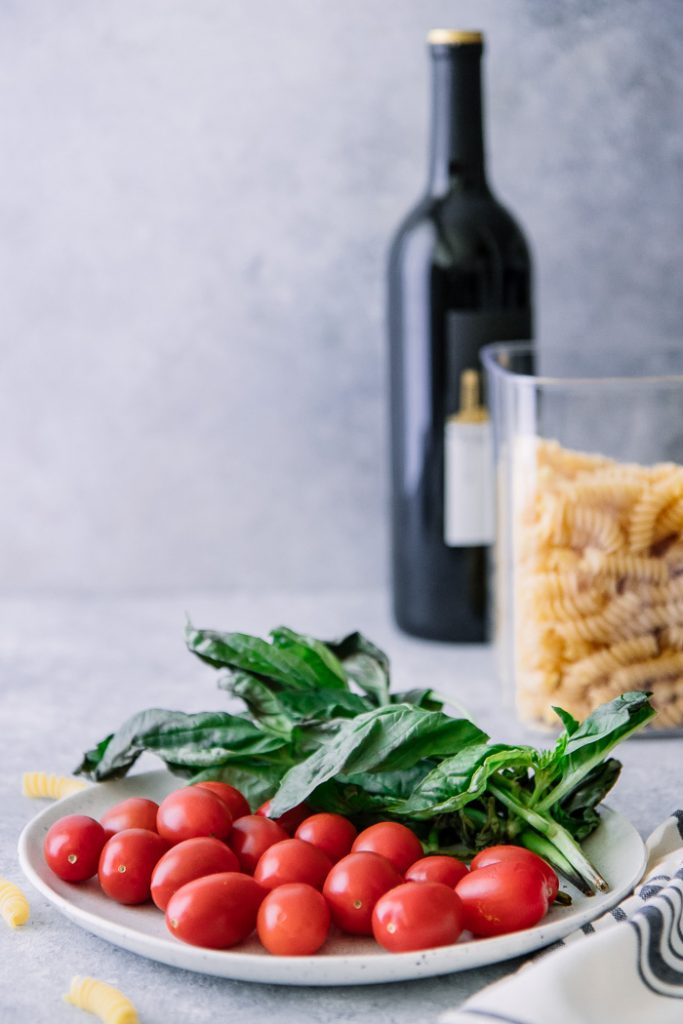 A white plate with cherry tomatoes and basil on a blue table with a jar of fusilli pasta and a bottle of wine in the background.