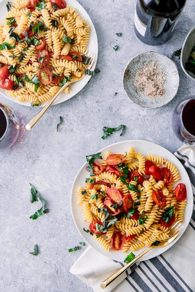 Two white plates of pasta salad with tomatoes, basil, and fusilli pasta on a blue table with a glass of wine.