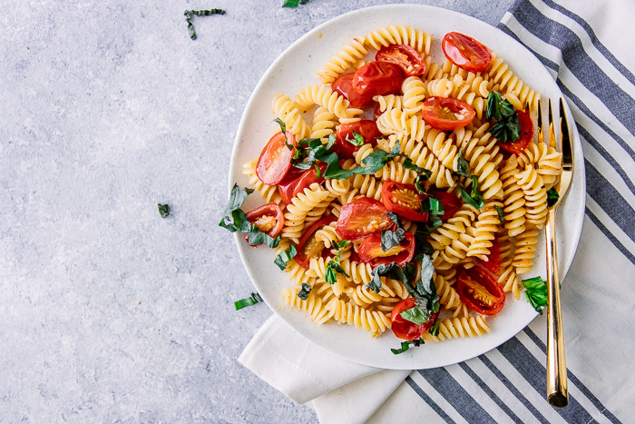 A white plate with fusilli garlic tomato basil pasta salad with a white napkin on a blue table.