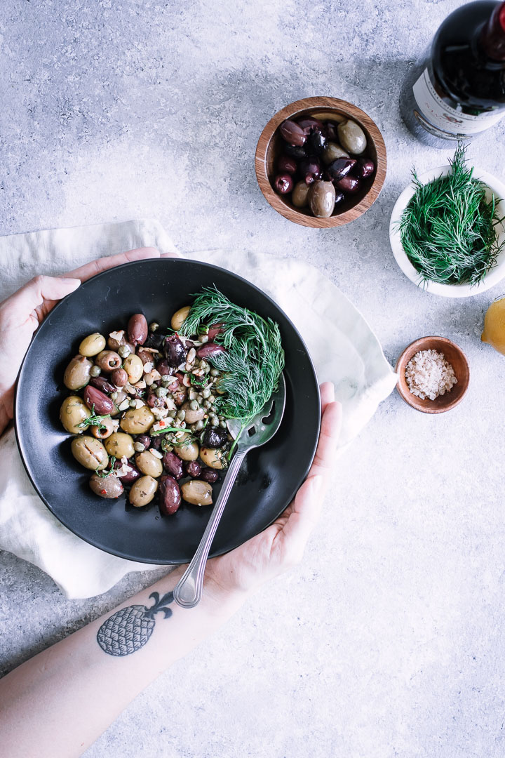 Two hands holding a black bowl of mixed olives on a blue table.