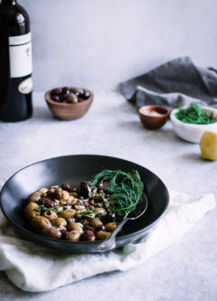 A black bowl with mixed olives and herbs with a bottle of wine and small bowls in the background.