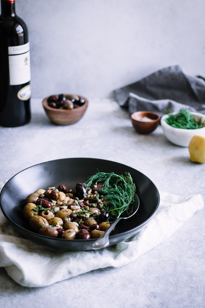 A black bowl with mixed olives and herbs with a bottle of wine and small bowls in the background.