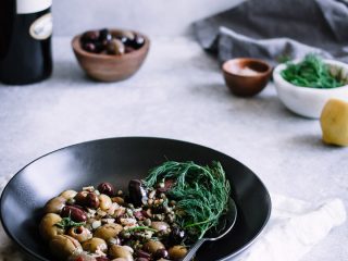 A black bowl with mixed olives and herbs with a bottle of wine and small bowls in the background with the words "warm marinated olives."