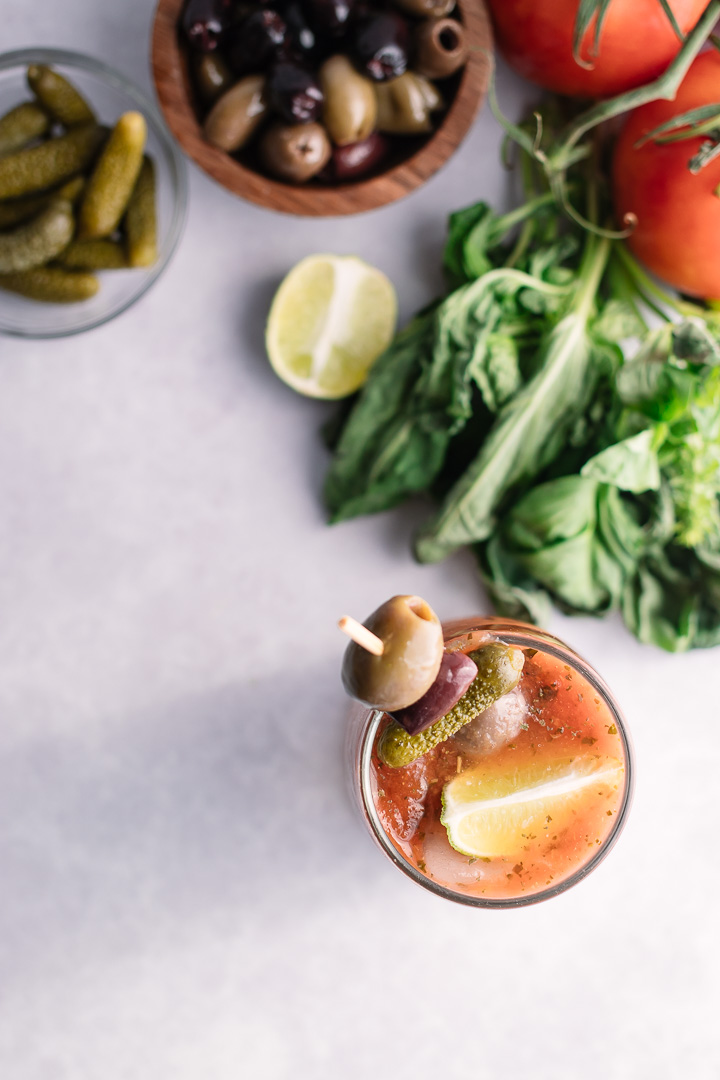 A flatlay photo of a bloody mary with olives and lime on a table with herbs and tomatoes.