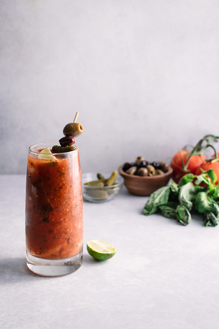 A bloody mary in a tall glass with olives, basil, pickles, and tomatoes.