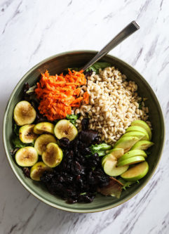 a fall grain bowl with warm barley, apples, figs, cranberries, carrots, and maple dressing in a green bowl on a white table