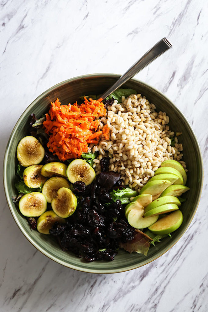 a fall grain bowl with warm barley, apples, figs, cranberries, carrots, and maple dressing in a green bowl on a white table