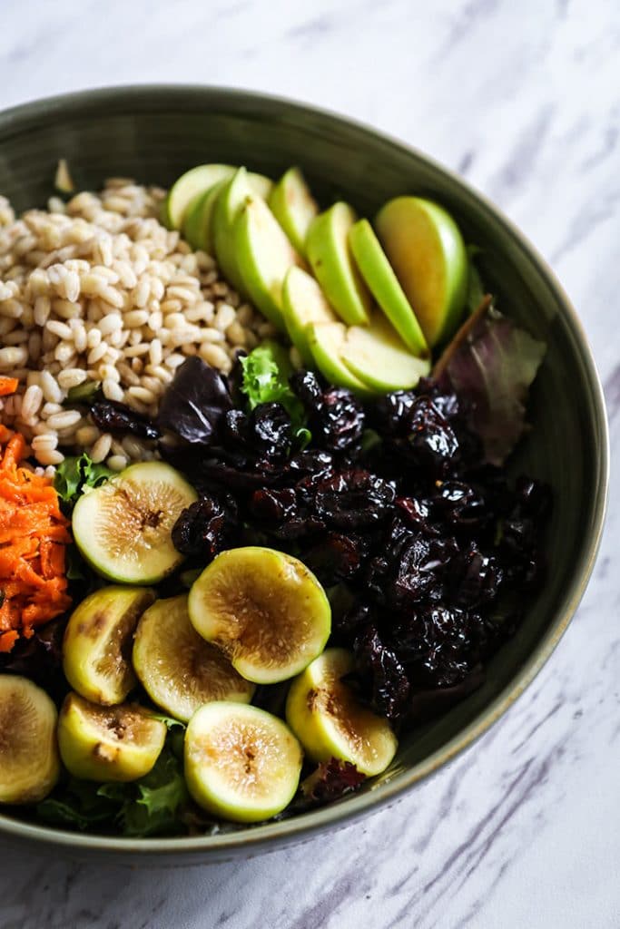 a green bowl filled with grains, sliced apples, sliced figs, dried cranberries, and shredded carrots on a white table