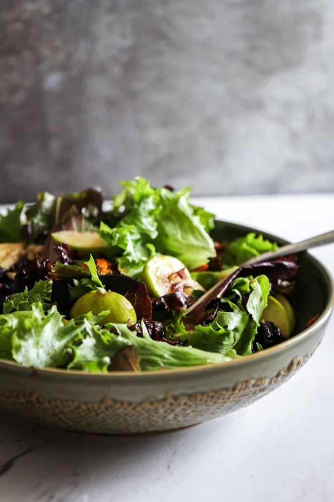 a large salad bowl with leafy greens, grains, and fall produce on a white table