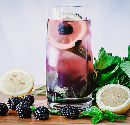 A glass of boozy blackberry lemonade on a cutting board with fresh fruit.