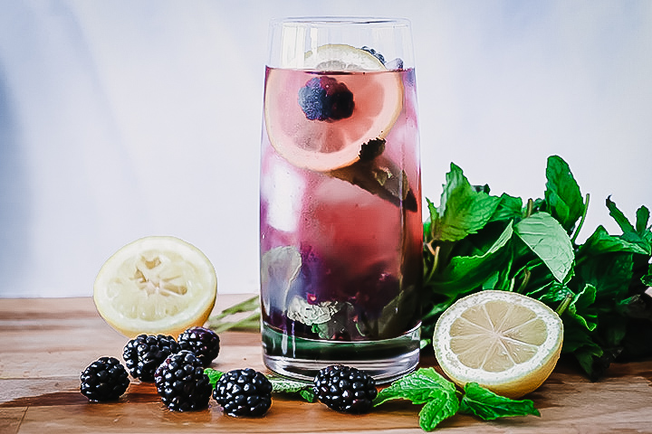 A glass of boozy blackberry lemonade on a cutting board with fresh fruit.