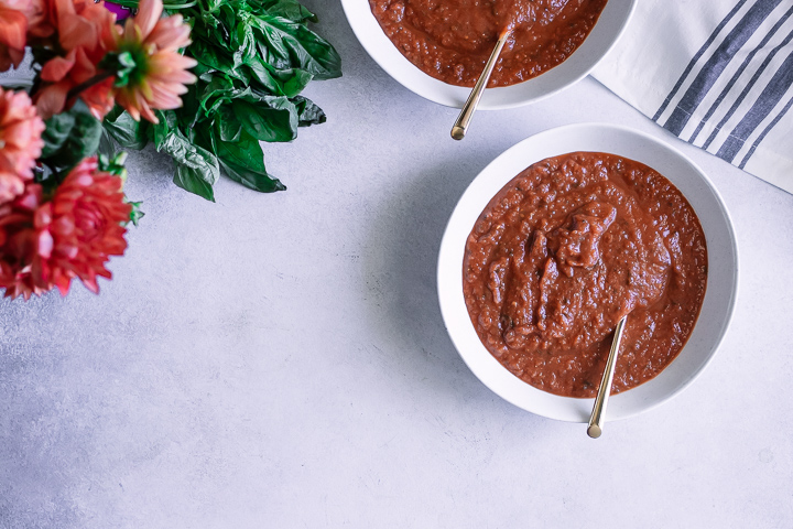 Two white soup bowls with red tomato sop on a white table with fresh basil.