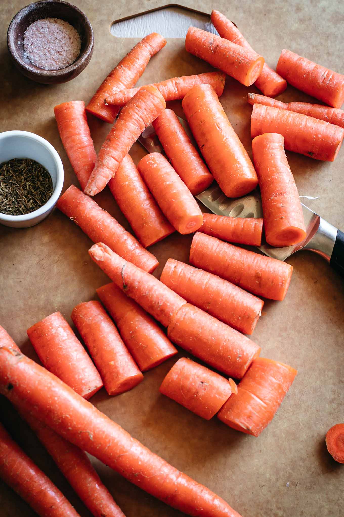 Chopped up carrots next to a knife on a cutting board, next to bowls of spices and salt