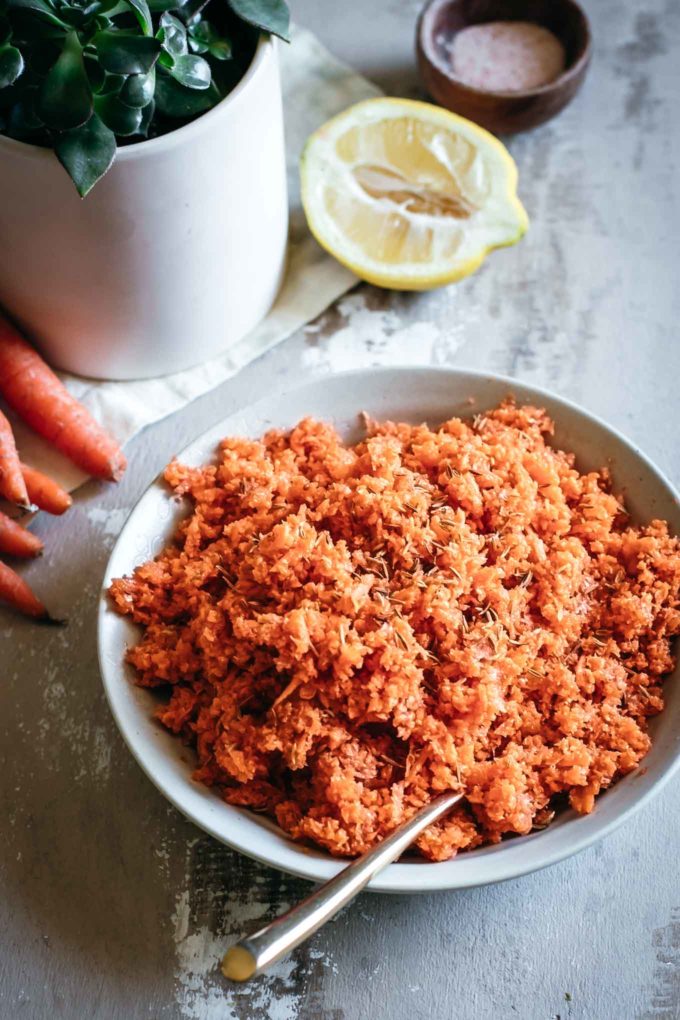 shredded carrot salad in a white bowl with a gold spoon, next to a cut lemon, a plant, and a bowl of salt