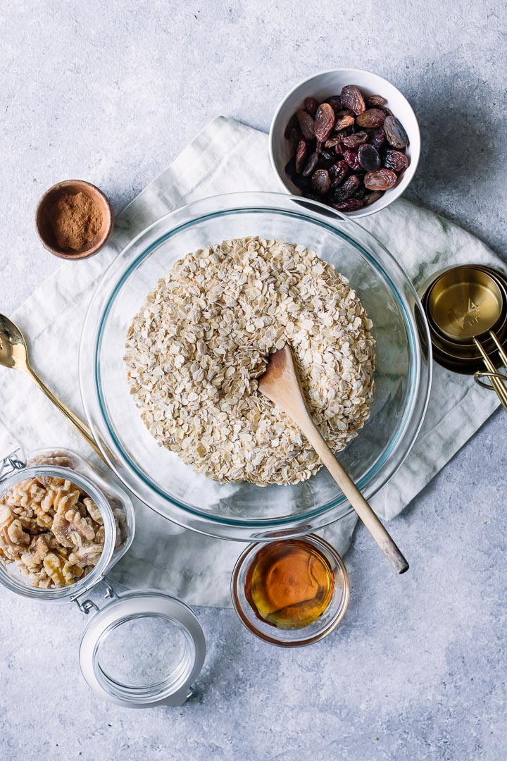 A mixing bowl with oats and small bowls of walnuts, cinnamon, maple syrup, and raisins on a white table.