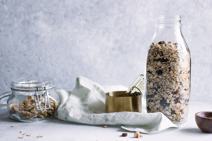 A jar of fresh homemade granola with a set of measuring cups and a white napkin on a white table.