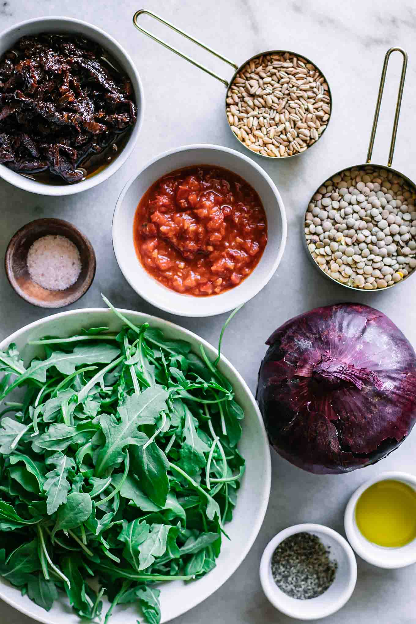 bowls of lentils, farro, arugula, onion, harissa, oil, salt, and pepper on a white table