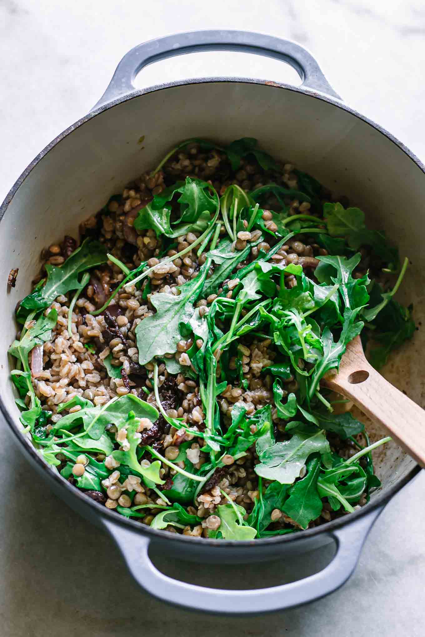 a large pot with lentils, farro, onion, sun-dried tomatoes, and arugula with a wooden spoon