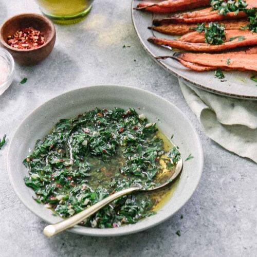 a bowl of green chimichurri sauce with carrot leaves in a blue bowl and a gold spoon