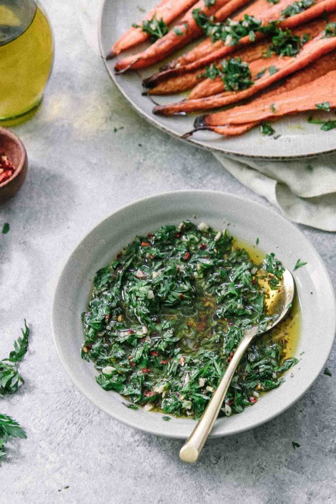 a bowl of green chimichurri sauce with carrot leaves in a blue bowl and a gold spoon