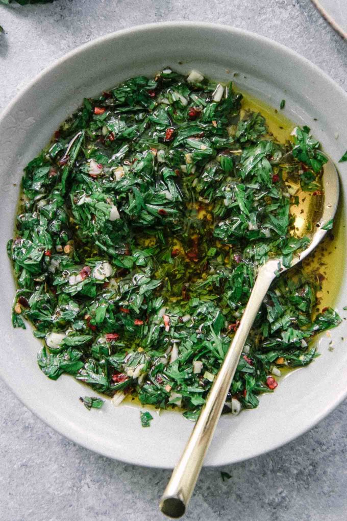 a close up photo of chimichurri in a blue bowl with a gold spoon