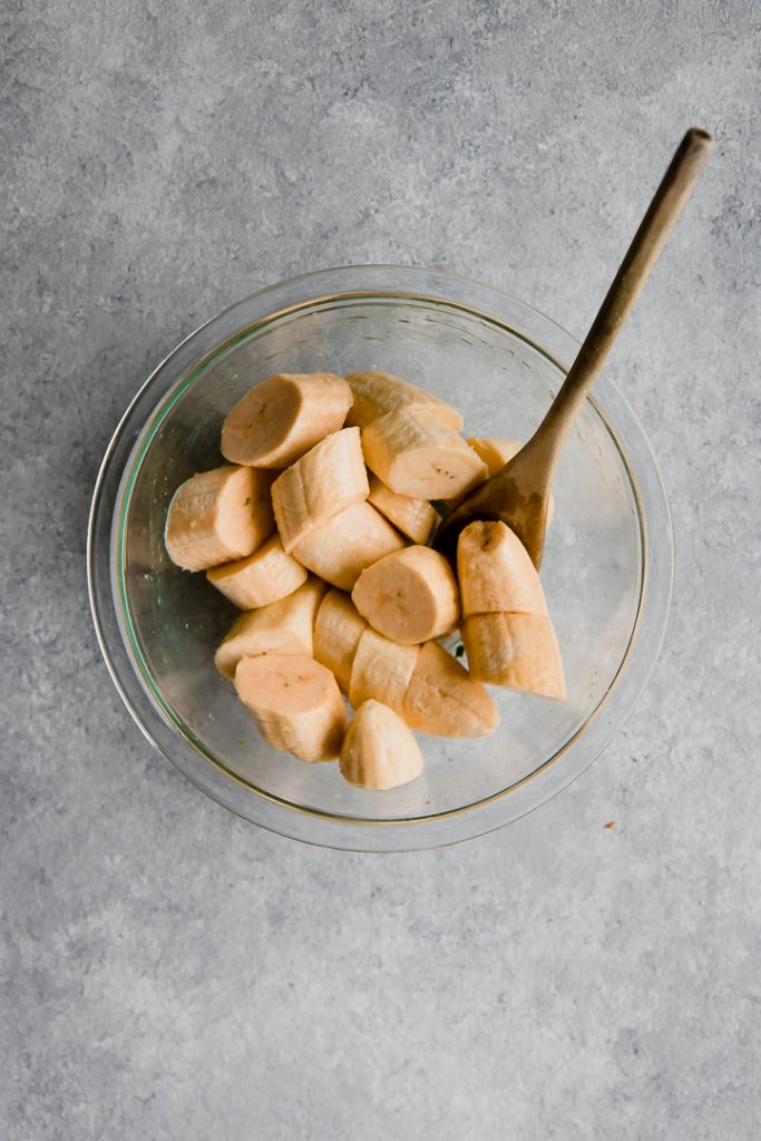 a clear glass bowl of sliced plantains with a wooden spoon for healthy baked patacones