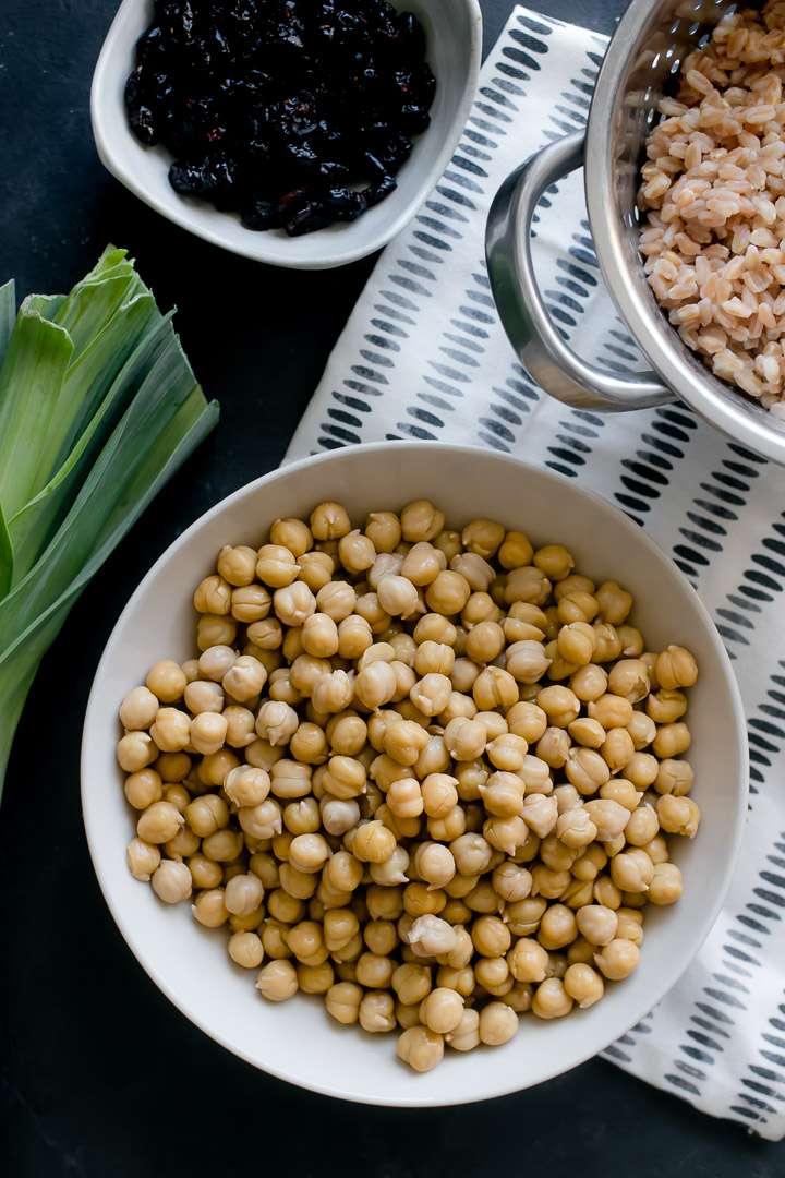 A bowl of chickpeas on a black table with a colander of farro and a leek on the side.