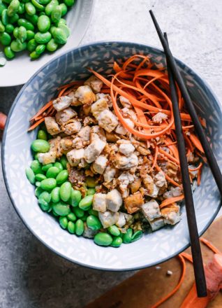 tofu, edamame, and shredded carrots over rice in a bowl with chopsticks on a white table