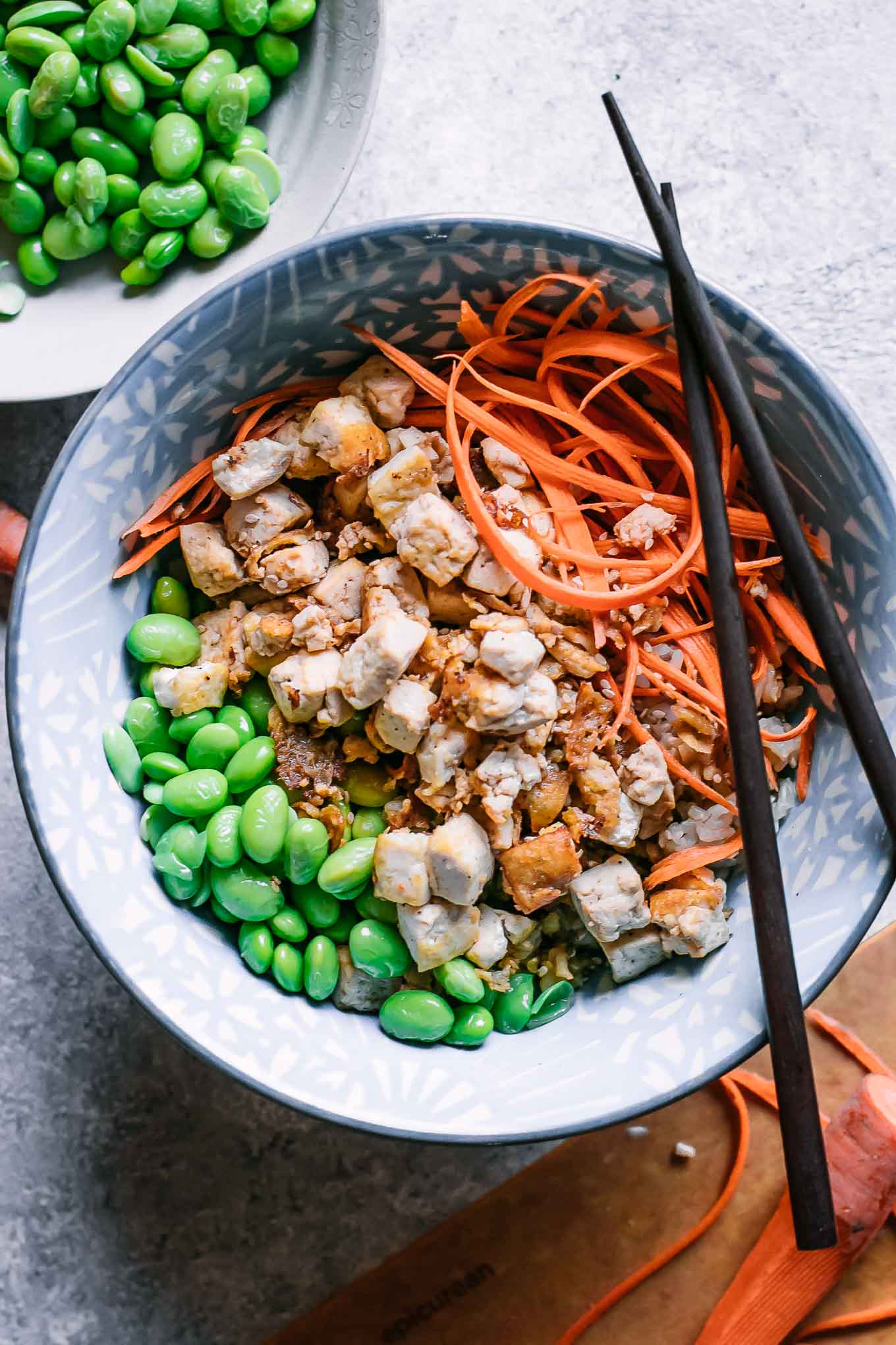 tofu, edamame, and shredded carrots over rice in a bowl with chopsticks on a white table