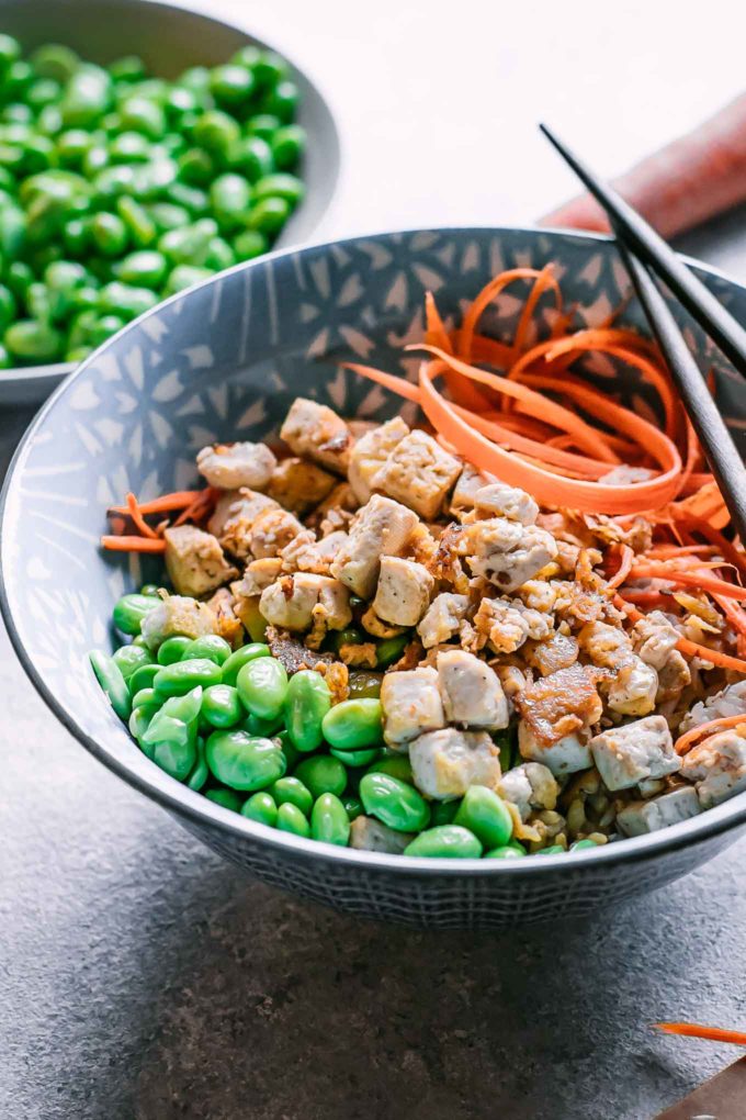 a tofu and rice bowl with carrots and edamame in a blue bowl on a white table