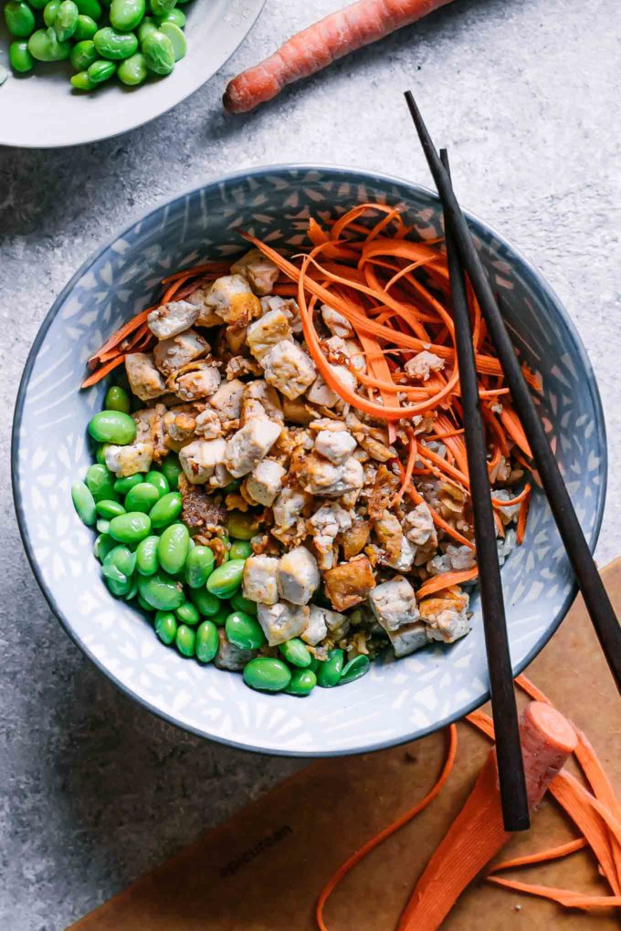 a rice and tofu bowl with edamame, carrots, and green onions on a white table