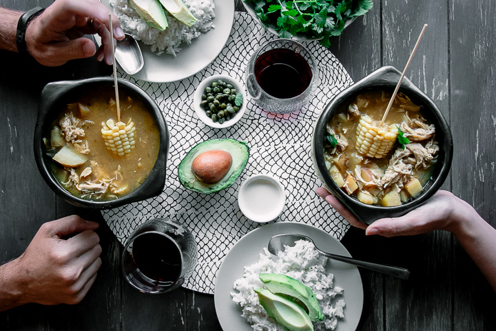 A chamba pot with Colombian ajiaco on a wood table with a side of rice, avocado, cream, and cilantro and a glass of wine and two people eating.