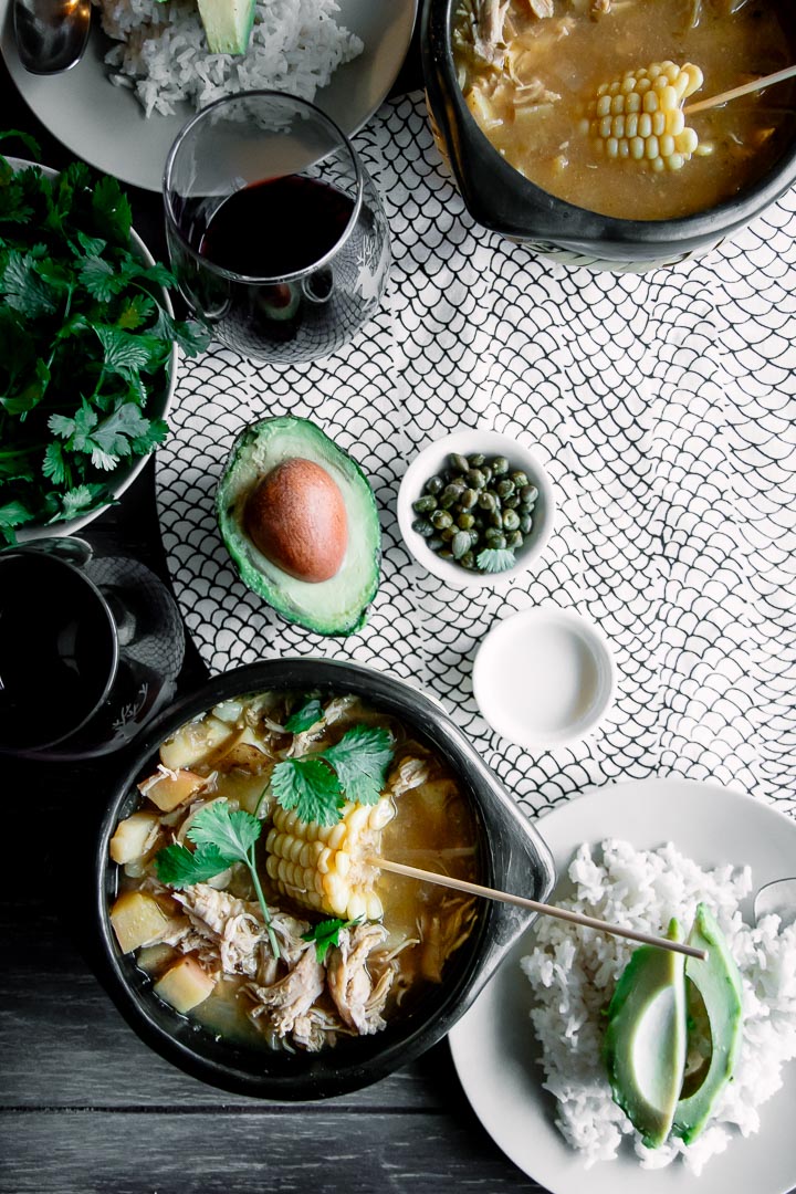 A chamba pot with Colombian ajiaco on a wood table with a side of rice, avocado, cream, and cilantro and a glass of wine.